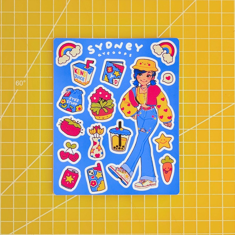 May include: A sticker sheet with a blue background featuring a cartoon girl wearing a yellow hat, a red jacket, and blue jeans. The sticker sheet also includes a rainbow, a juice box, a flower pot, a boba tea, a can of soda, and other cute illustrations. The text "Sydney aveoops" is at the top of the sheet.