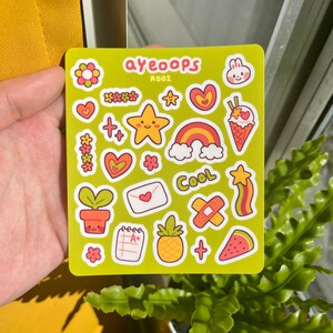 May include: A sheet of green sticker paper with a variety of colorful stickers, including a rainbow, a star, a pineapple, a watermelon, a plant, a heart, a bunny, a flower, a plus sign, a letter, a notepad, a bandage, and the text "ayeoops A002" and "Cool".