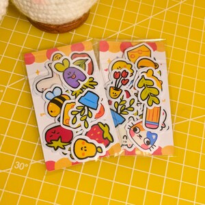 May include: Two sheets of colourful stickers featuring cute cartoon characters and objects. The stickers include a bee, a strawberry, a pear, a cat, a flower, a pencil, and a leaf.