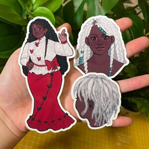 May include: Three stickers featuring illustrations of Black women. One sticker shows a woman in a red skirt and white blouse with butterflies. Another shows a woman with white braids and turquoise beads. The third shows the back of a woman's head with white braids.