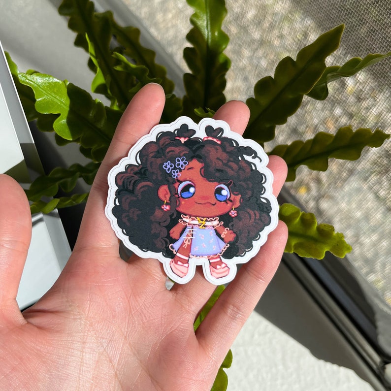 K&ouml;nnte beinhalten: Ein Aufkleber mit einer Cartoon-Illustration eines M&auml;dchens mit braunen Locken, das ein blau-wei&szlig;es Kleid mit rosa Blumen tr&auml;gt. Sie hat blaue Augen, rosafarbene Wangen und tr&auml;gt rosa Ohrringe.