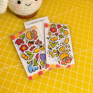 May include: Two packs of colourful flower stickers with a white background. The stickers feature various flowers, plants, and smiling faces. The text "Flowers Only" is printed on the top of one of the packs.