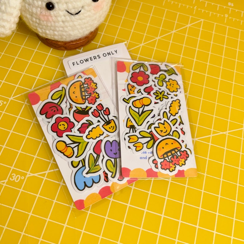 May include: Two packs of colourful flower stickers with a white background. The stickers feature various flowers, plants, and smiling faces. The text "Flowers Only" is printed on the top of one of the packs.