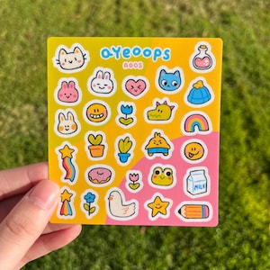 May include: A sheet of colorful stickers with various cute illustrations, including cats, bunnies, hearts, and a rainbow. The stickers are on a yellow background with the text "ayeoops" in blue. The stickers are held by a hand.