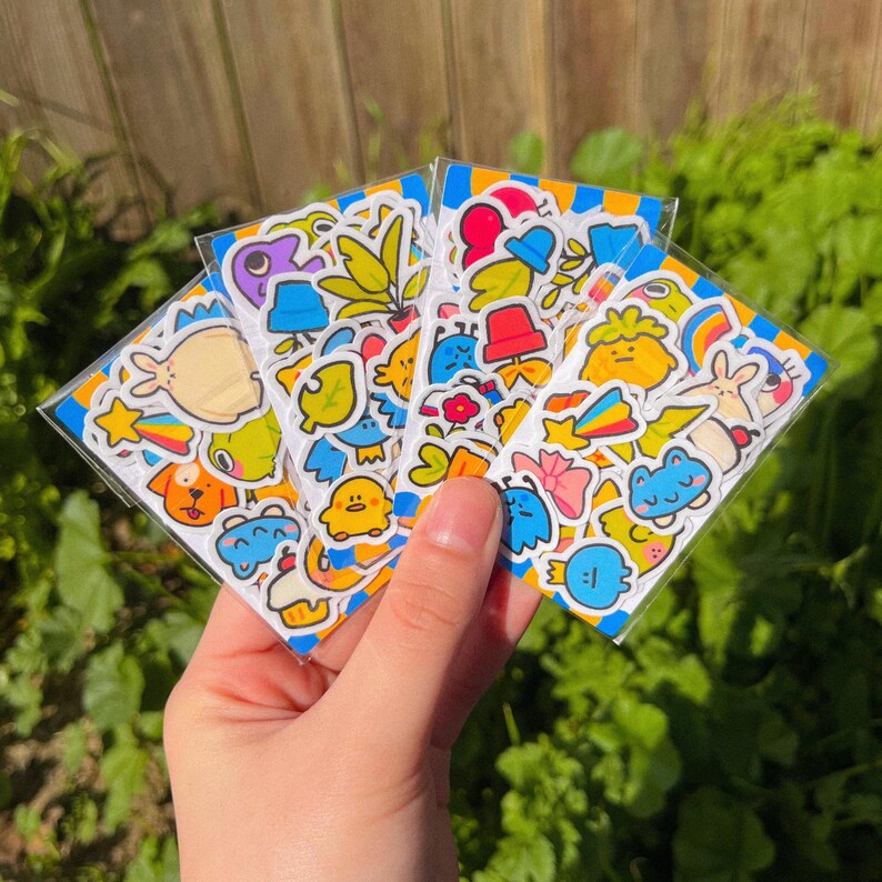 May include: A set of colourful stickers featuring cute cartoon characters, including animals, plants, and other whimsical designs. The stickers are printed on white paper and have a glossy finish.
