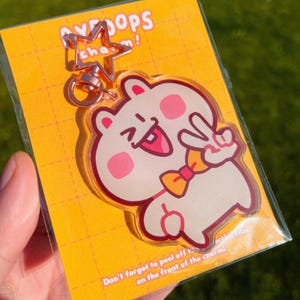 May include: A clear acrylic keychain featuring a cartoon character with a pink nose, a yellow bow tie, and a peace sign. The keychain is attached to a silver star charm and a silver chain.
