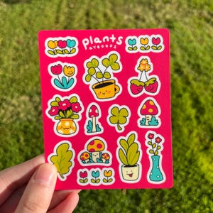 May include: A pink sheet of stickers with the words "Plants Ayeoops" at the top. The stickers feature various plants, flowers, and mushrooms in a cartoon style. There are red, yellow, blue, green, and brown colors used in the stickers.