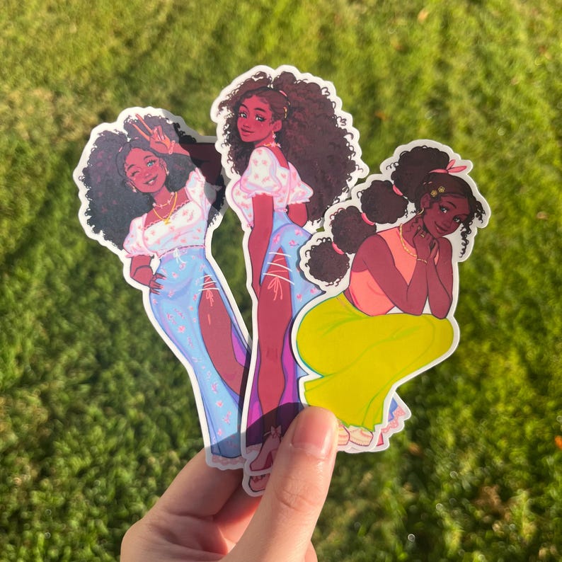 May include: Three colorful stickers featuring illustrations of women with brown skin and curly hair. The women are wearing different outfits, including a blue dress with white flowers, a white dress with pink flowers, and a yellow skirt with a pink top.