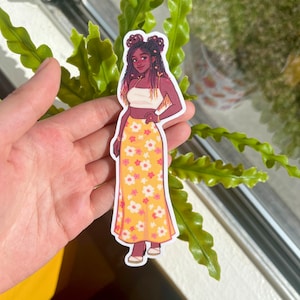 May include: A sticker of a woman with brown skin, wearing a white crop top and a yellow skirt with pink flowers. She has brown hair with two buns and a gold bracelet on her wrist.