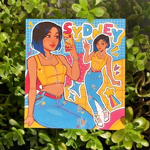 May include: A digital illustration of a woman with blue hair wearing a yellow crop top, blue jeans with stars and a heart, and a necklace. She is holding a phone and smiling. The background is a blue and yellow grid with the word "Sydney" in large letters.