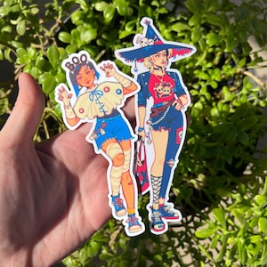 May include: Two illustrated stickers of women dressed in Halloween costumes. One sticker features a woman with a witch hat, black and red outfit, and a skull and crossbones design. The other sticker features a woman with a blue and white outfit and the word "BOO" on her hat.