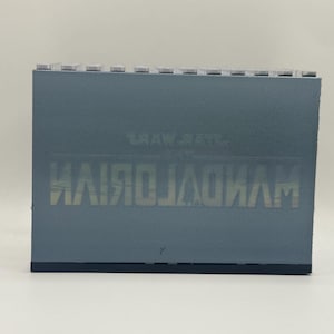 May include: A rectangular, gray-blue object with the text "THE MANDALORIAN" printed in a mirrored, light-colored font. The top edge has a series of small, light-colored rectangular protrusions. The object is set against a plain white background.