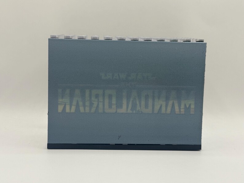 May include: A rectangular, gray-blue object with the text "THE MANDALORIAN" printed in a mirrored, light-colored font. The top edge has a series of small, light-colored rectangular protrusions. The object is set against a plain white background.