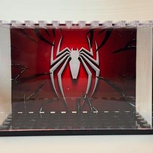 May include: A clear acrylic display case with a black base featuring a series of studs. The interior back panel displays a red background with a large white spider emblem and black tendril-like graphics extending from the edges toward the center.