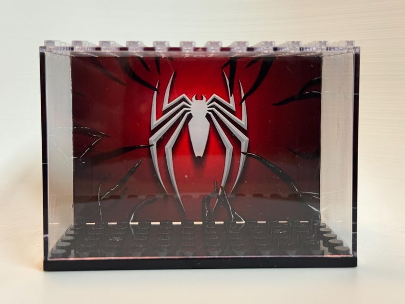 May include: A clear acrylic display case with a black base featuring a series of studs. The interior back panel displays a red background with a large white spider emblem and black tendril-like graphics extending from the edges toward the center.