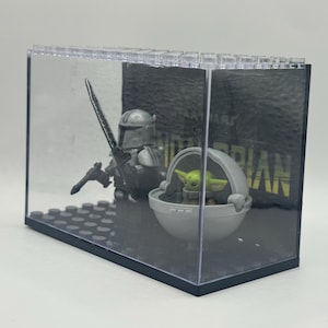 May include: A clear display case featuring a Mandalorian figure with a blaster and a green-skinned Grogu figure in a floating pod. The background has the word "MANDALORIAN" in white letters on a dark gray surface. The case is made of clear plastic.
