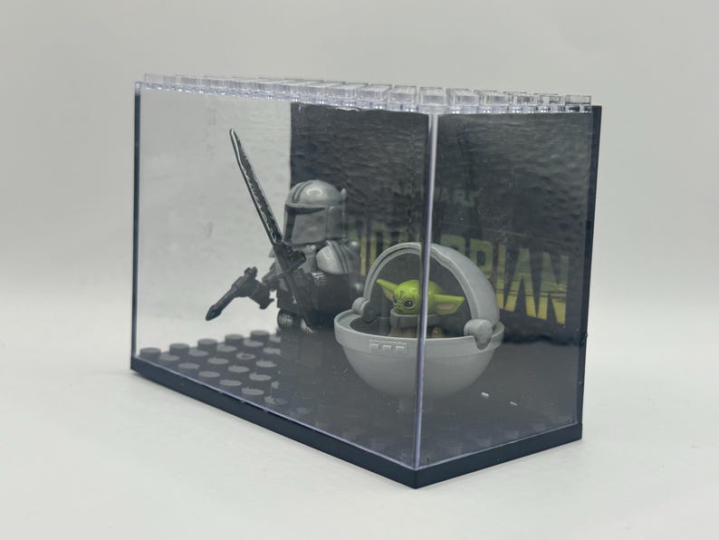 May include: A clear display case featuring a Mandalorian figure with a blaster and a green-skinned Grogu figure in a floating pod. The background has the word "MANDALORIAN" in white letters on a dark gray surface. The case is made of clear plastic.