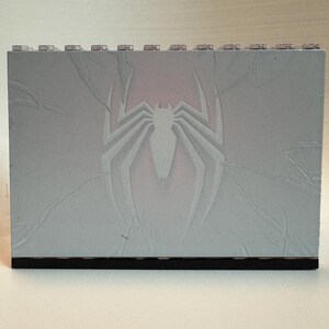 May include: A rectangular light gray building block panel featuring a debossed spider emblem in the center. The surface displays subtle crack-like textures radiating from the spider icon, and the top edge has a row of small circular studs.