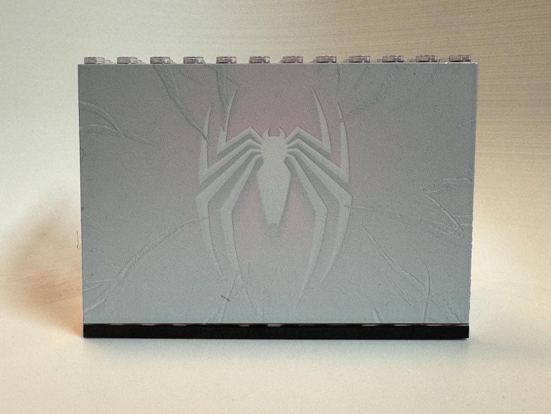 May include: A rectangular light gray building block panel featuring a debossed spider emblem in the center. The surface displays subtle crack-like textures radiating from the spider icon, and the top edge has a row of small circular studs.