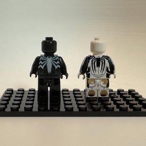 May include: Two plastic minifigures standing on a black rectangular building block base. The left figure is black with a white spider emblem on the chest. The right figure is white with a black spider emblem on the chest and black arm details.