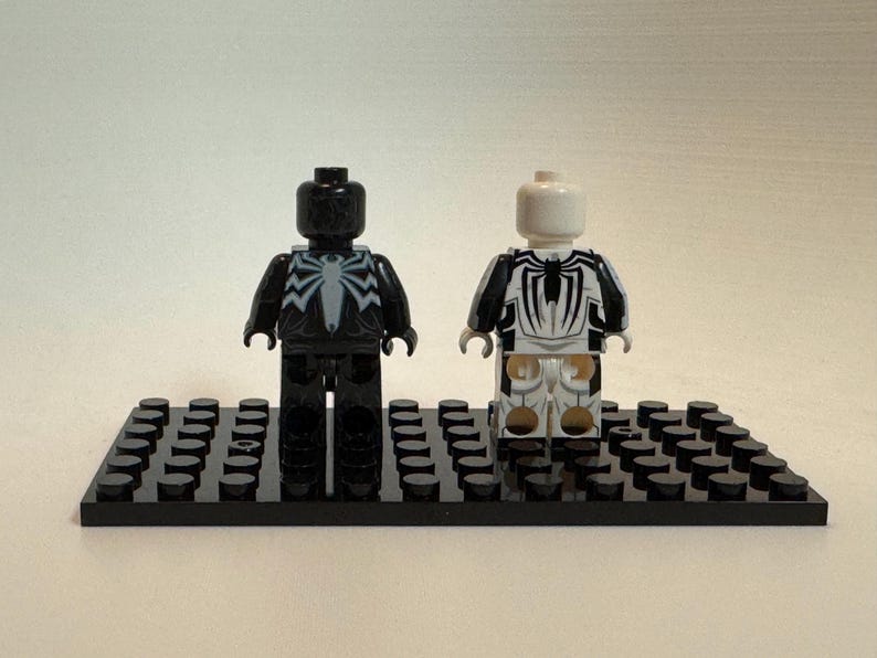 May include: Two plastic minifigures standing on a black rectangular building block base. The left figure is black with a white spider emblem on the chest. The right figure is white with a black spider emblem on the chest and black arm details.