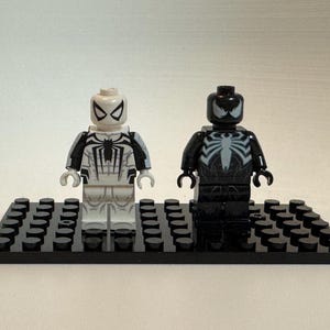May include: Two plastic minifigures standing on a black rectangular building block base. The left figure is white with black spider web patterns and a black spider emblem. The right figure is black with a large white spider emblem on the chest.