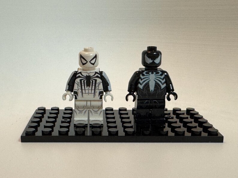 May include: Two plastic minifigures standing on a black rectangular building block base. The left figure is white with black spider web patterns and a black spider emblem. The right figure is black with a large white spider emblem on the chest.