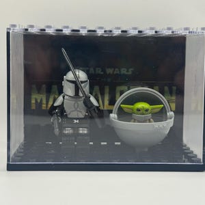 May include: A clear display case featuring Lego figures of The Mandalorian and Grogu from Star Wars. The Mandalorian figure is in silver armor, holding a weapon. Grogu is in a gray floating pod. The background reads "STAR WARS THE MANDALORIAN."