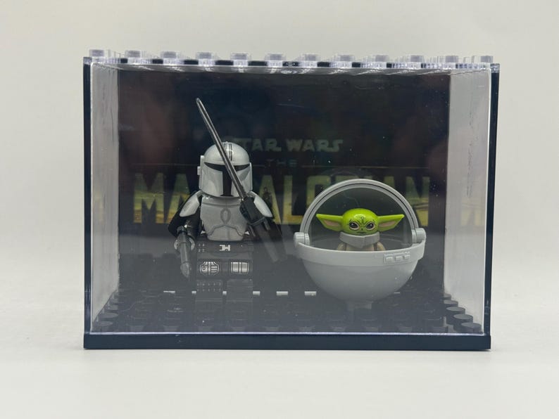 May include: A clear display case featuring Lego figures of The Mandalorian and Grogu from Star Wars. The Mandalorian figure is in silver armor, holding a weapon. Grogu is in a gray floating pod. The background reads "STAR WARS THE MANDALORIAN."