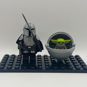 May include: A Lego set featuring The Mandalorian figure with a blaster and a green-skinned Grogu figure in a floating pram. The figures are on a black baseplate. The Mandalorian is in silver armor.