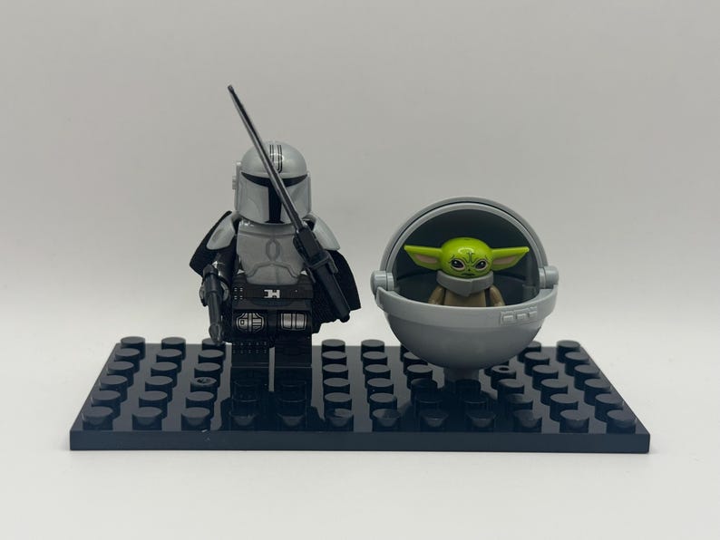 May include: A Lego set featuring The Mandalorian figure with a blaster and a green-skinned Grogu figure in a floating pram. The figures are on a black baseplate. The Mandalorian is in silver armor.