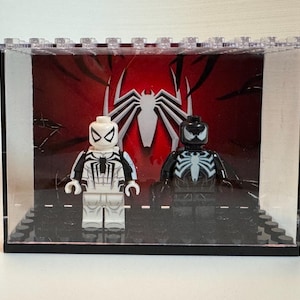 May include: Two plastic minifigures displayed inside a clear rectangular case. One figure is white with black spider-themed details, and the other is black with white spider-themed details. The background features a large white spider emblem on red.