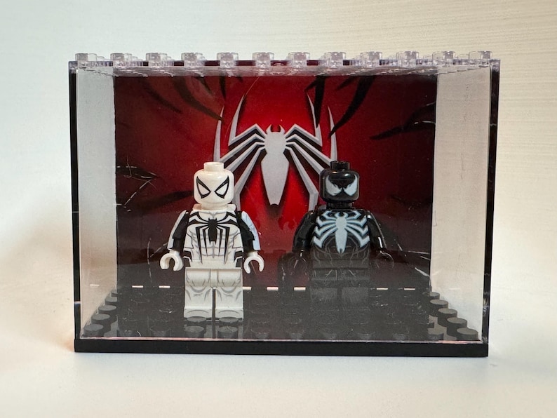 May include: Two plastic minifigures displayed inside a clear rectangular case. One figure is white with black spider-themed details, and the other is black with white spider-themed details. The background features a large white spider emblem on red.