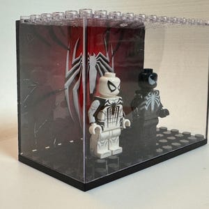 May include: A clear acrylic display case containing two plastic minifigures standing on a black studded base. One figure is white with black spider-themed markings, and the other is black with a white spider emblem. A red and black backdrop features a large white spider logo.