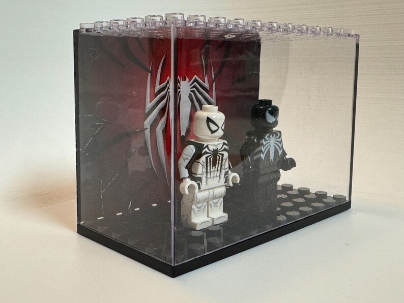 May include: A clear acrylic display case containing two plastic minifigures standing on a black studded base. One figure is white with black spider-themed markings, and the other is black with a white spider emblem. A red and black backdrop features a large white spider logo.
