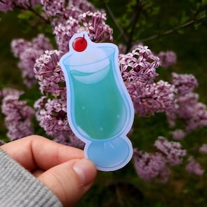 May include: A sticker featuring a cartoon-style blue cocktail with a red cherry on top. The drink is in a tall glass with a rounded base. The sticker is held in front of a background of purple flowers.