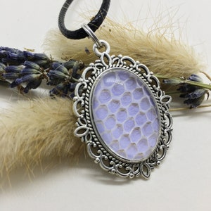 Op de afbeelding: Ovale hangerketting met een zilverkleurig filigraan frame. De hanger heeft een lichtpaars en wit honingraatpatroon. De ketting heeft een zwart leren koord.