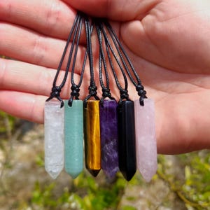 May include: A collection of seven crystal pendant necklaces in various colors, including clear, green, yellow, purple, black, and pink. Each crystal is rectangular with a pointed end and is suspended from a black cord.