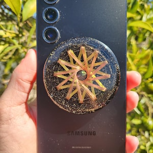 以下が含まれることがあります： ゴールドと銅の電話グリップが付いた黒いサムスンスマートフォン。グリップは、星形の模様が施された円形のデザインです。