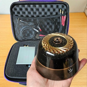 May include: A black and gold audio device with a spiral design is held in front of an open purple case containing audio equipment. The device has a copper-colored base and a gold spiral top. The case holds a silver component, cables, and connectors.