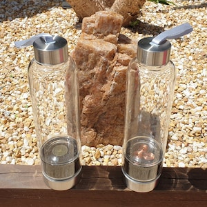 Peut inclure: Deux bouteilles d'eau en verre transparent avec des couvercles argentés et des poignées en tissu gris. Les bouteilles contiennent une bobine de cuivre à l'intérieur.