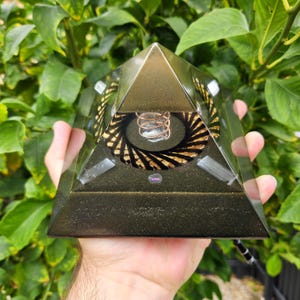 Könnte beinhalten: Ein dunkelgrünes pyramidenförmiges Objekt mit goldfarbener Spitze und einem Spiraldesign in der Mitte. Die transparente Pyramide zeigt interne Komponenten und eine Kupferspule. Das Objekt wird in einer Hand gehalten, vor einem Hintergrund aus grünen Blättern. Ca. 10 cm.