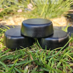 May include: Three black circular soap bars stacked on top of each other on a bed of green grass.