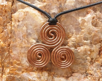 May include: A copper-colored pendant featuring a triple spiral design, suspended from a black cord. The pendant's spirals are tightly wound, creating a symmetrical pattern. The necklace rests against a textured, light-colored stone surface.