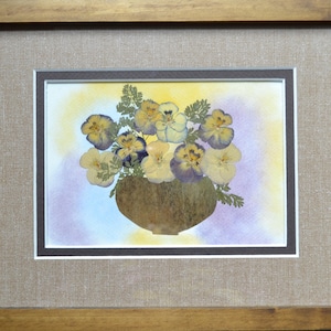 Puede incluir: Una obra de arte enmarcada que presenta un ramo de pensamientos prensados en un jarrón. Los pensamientos son en tonos de amarillo, blanco y morado, sobre un fondo de acuarela. La obra está enmarcada en un marco de madera con un paspartú con textura de lino.