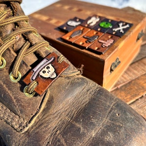May include: A brown leather boot with a decorative leather tag featuring a skull wearing a hat. A wooden box with several small leather squares with various designs is in the background. The boot has brown laces and brass eyelets.