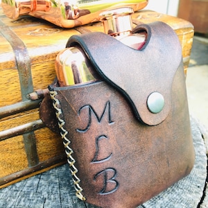 May include: A copper flask and lid sit on a wooden chest. A brown leather flask holder with the initials "MLB" is in the foreground. The holder is stitched with white thread and has a snap closure. The flask is a stylish accessory.