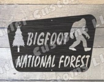 Może przedstawiać: Czarny metalowy znak z sylwetką Bigfoota idącego w prawo. Znak zawiera napis "Bigfoot National Forest" oraz grafikę sosny po lewej stronie.