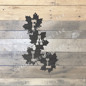 Könnte beinhalten: Schwarzer Metallausschnitt des Wortes "FALL" mit jedem Buchstaben in einer Ahornblattform. Die Blätter sind übereinander gelegt. Der Hintergrund ist hellbraunes Holz.
