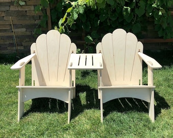 May include: Two white wooden chairs with a scalloped back and a small table between them. The chairs and table are on a green grassy lawn.